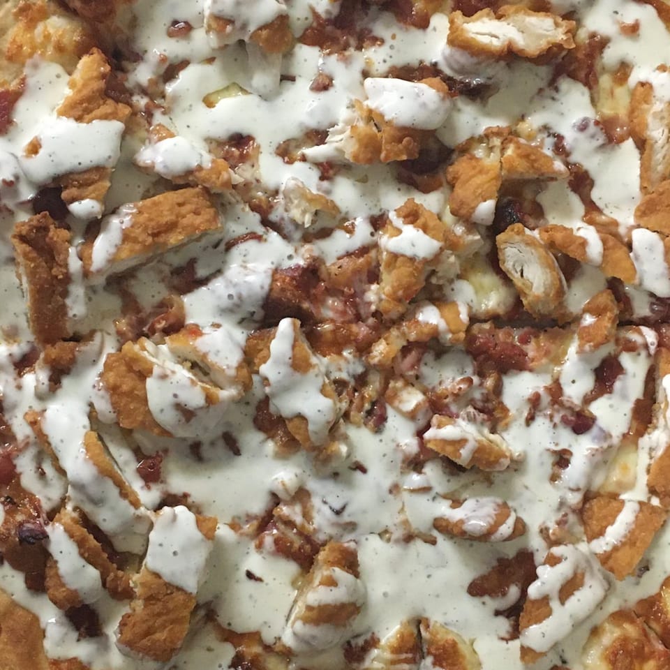 Close-up of a pizza topped with crispy chicken pieces, bacon, and a drizzle of white sauce.