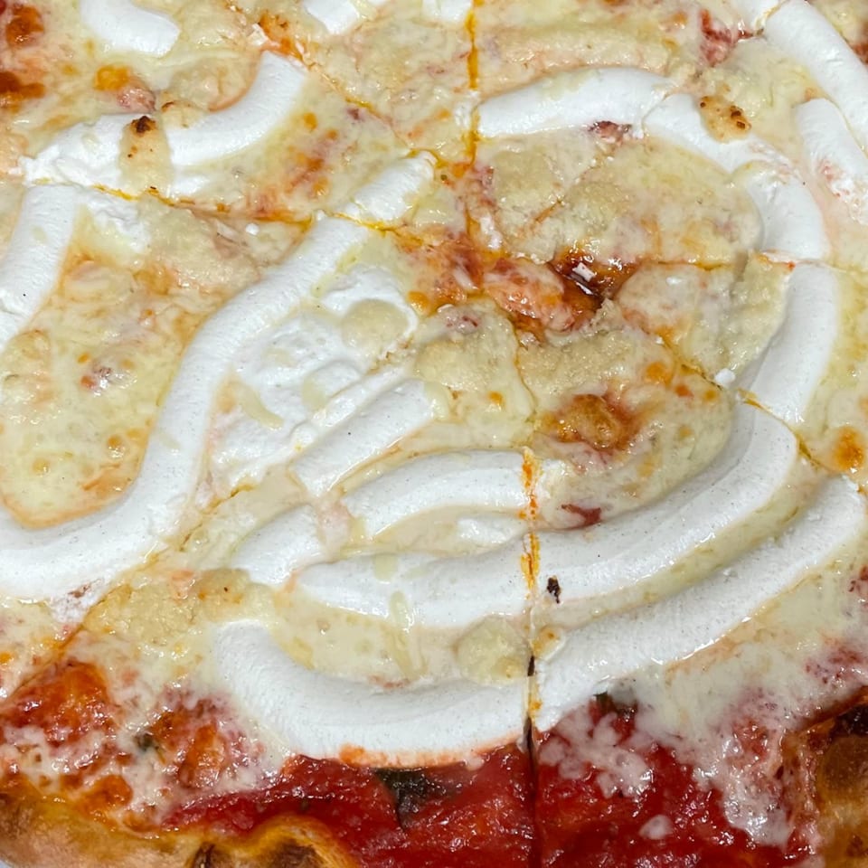 Close-up of a sliced Amici Vicinato pizza with melted cheese, red sauce, and white cream swirls.